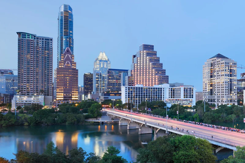 The Austin, TX skyline at dusk, viewed across the water with a pedestrian bridge in the foreground