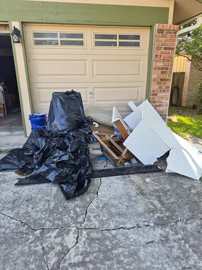 Black trash bags, white panels, and broken wood piled on the driveway in front of a garage door before removal