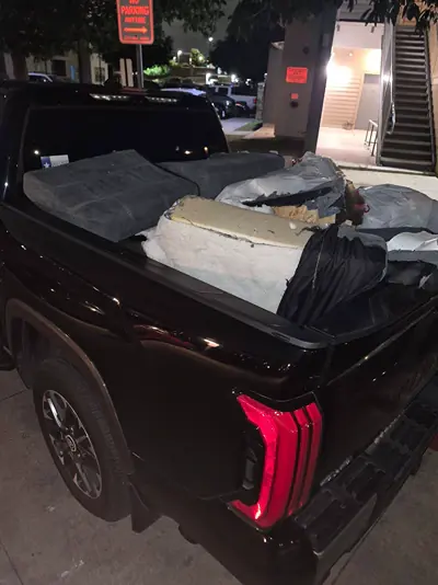 Couch cushions, a mattress, and other bulky items loaded in the bed of a black pickup truck at night outside an apartment building