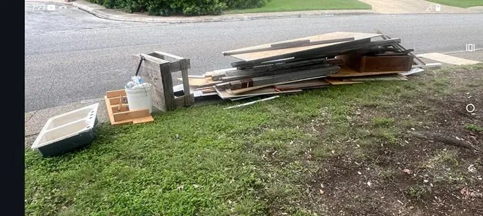 Old lumber, a wooden stool, a white bucket, and a plastic basin piled on the grass beside the curb before pickup