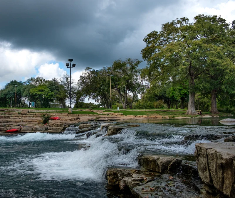 The San Marcos River flowing over a rock spillway, with storm clouds overhead