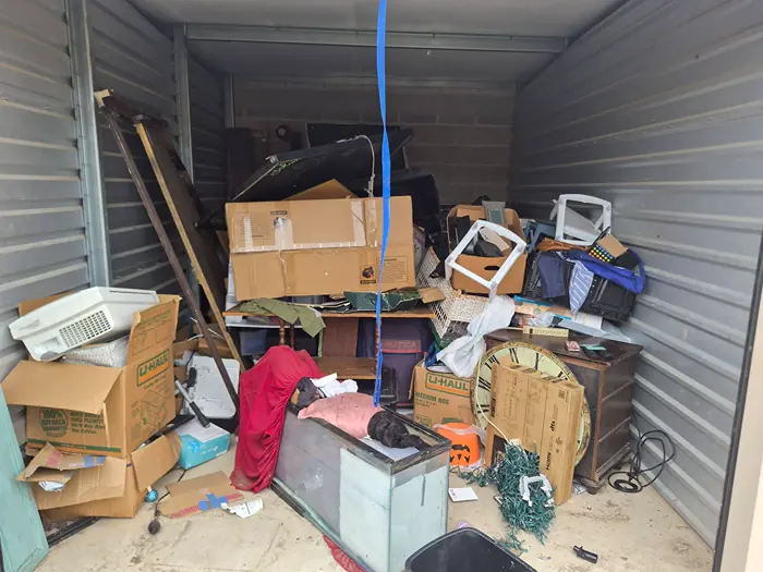Storage unit crowded with boxes, a wooden cabinet, a fish tank, furniture, and other household items before the cleanout